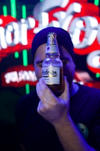 a man holding up a bottle of beer in front of a neon sign